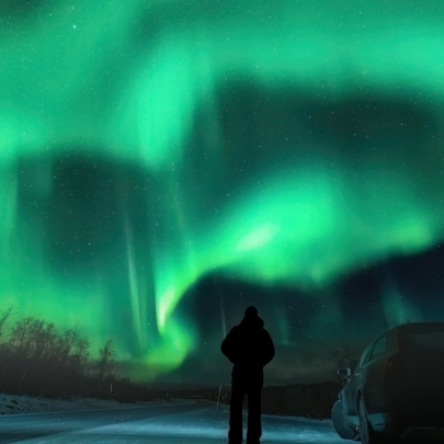 Homme arrêté près d'une voiture sur une route enneigée sous un ciel illuminé par les aurores boréales, lors d’une soirée de chasse aux lumières à Muonio